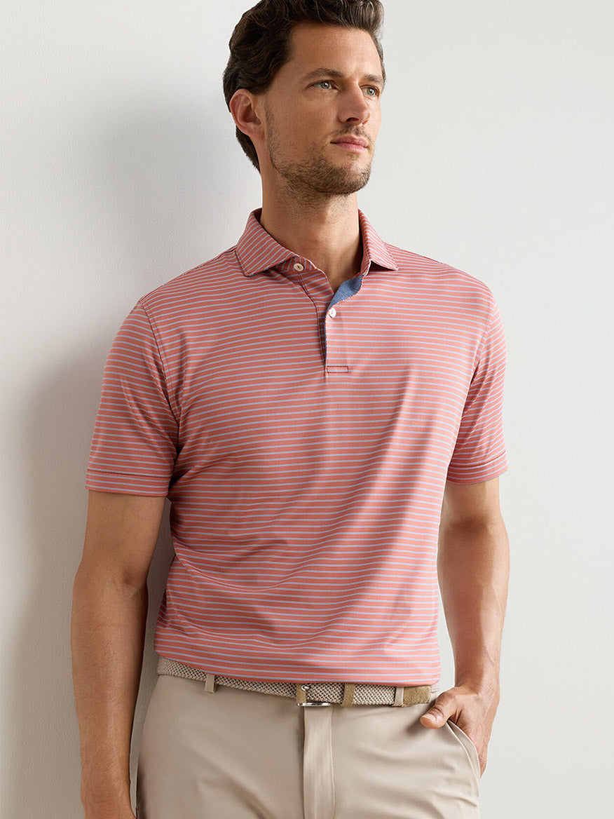 A man stands against a light background wearing the Peter Millar Keynote Performance Jersey Polo in Faded Scarlet, paired with beige pants and a woven belt.