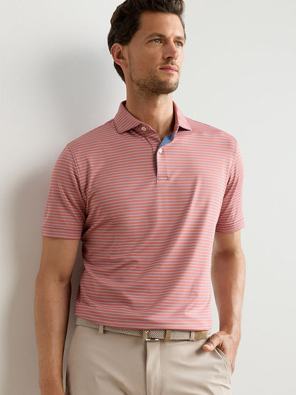 A man stands against a light background wearing the Peter Millar Keynote Performance Jersey Polo in Faded Scarlet, paired with beige pants and a woven belt.