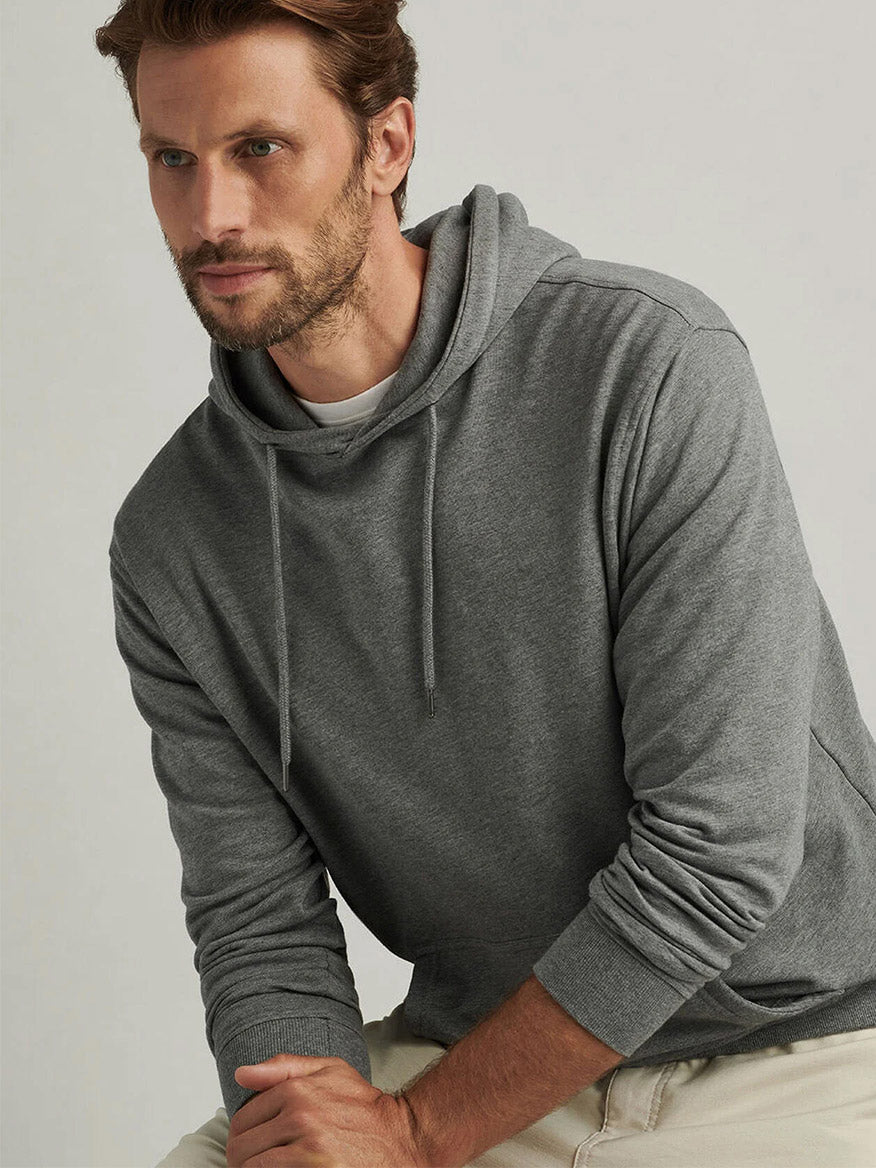 A man with light brown hair and a beard wears the Peter Millar Lava Wash Hoodie in Gale Grey, made from super-soft cotton-modal for ultimate comfort, paired with light-colored pants as he sits against a plain background.