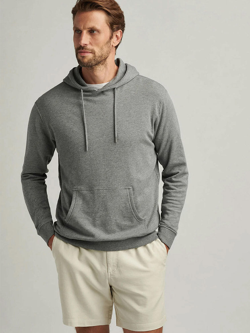 A man stands with hands in pockets, wearing the Peter Millar Lava Wash Hoodie in Gale Grey and beige shorts against a plain background, highlighting the hoodie’s super-soft comfort and effortless style.