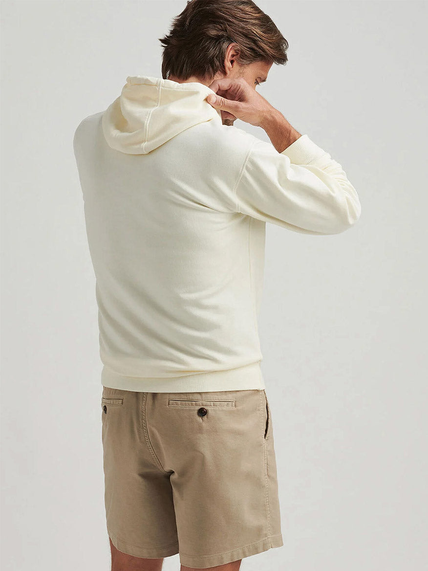 A person faces away, adjusting the hood of a Peter Millar Lava Wash Hoodie in Salt Water Taffy—a super-soft, classic fit cream-colored style—paired with beige shorts against a plain light background.