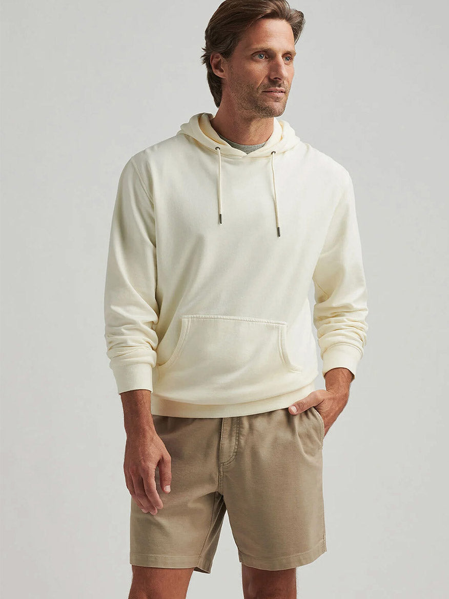 A man wearing the Peter Millar Lava Wash Hoodie in Salt Water Taffy and beige shorts stands against a light background, embodying all-day comfort and a classic fit with one hand in his pocket.