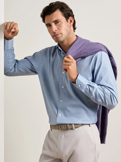 A man leans against a white wall and looks at the camera, wearing the Peter Millar Liam Crown Soft Twill Sport Shirt in Blue Freesia—crafted from easy-care, stretch performance fabric—with a purple sweater draped over his shoulders.