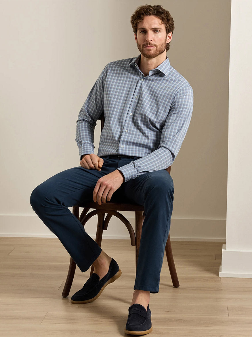 A man with short brown hair and a beard sits on a wooden chair in a minimalist room with light wood flooring, wearing the Peter Millar Lincoln Crown Lite Cotton-Stretch Sport Shirt in Navy, classic-fit navy pants, and dark loafers.