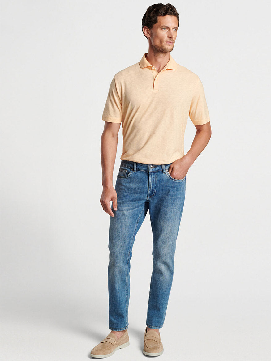 Man wearing a beige Peter Millar polo shirt and Peter Millar Vintage Washed Five-Pocket Denim in Stone Washed Blue standing against a neutral background.