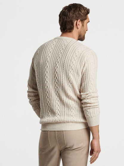 A man with light brown hair is wearing the Peter Millar Crescent Ridge Cable Crew in Winter Ivory, a cream-colored cable knit sweater made from Merino wool, along with beige pants. Perfect for 3-season wear, he stands with his back to the camera.