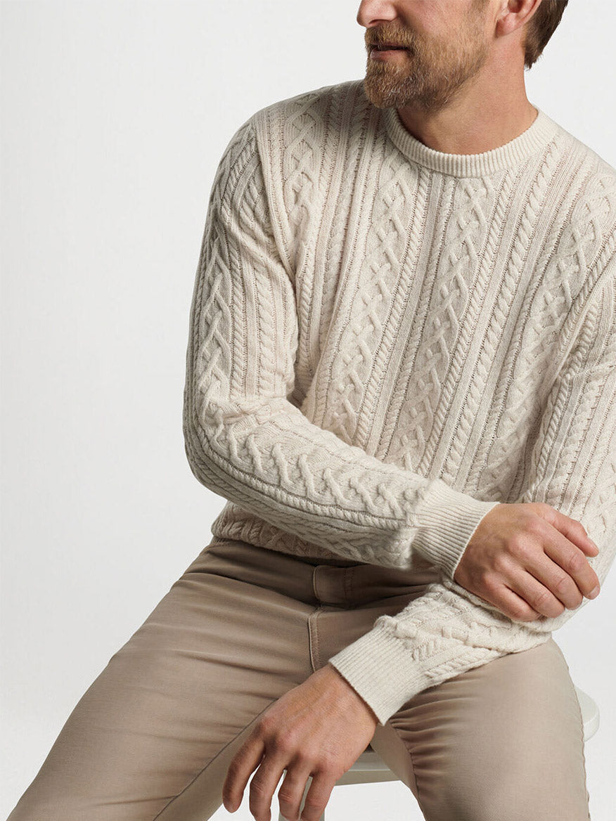 Set against a plain backdrop, the person is wearing the Peter Millar Crescent Ridge Cable Crew in Winter Ivory, a cream cable-knit sweater made from Merino wool that’s ideal for three-season wear, complemented by beige pants.