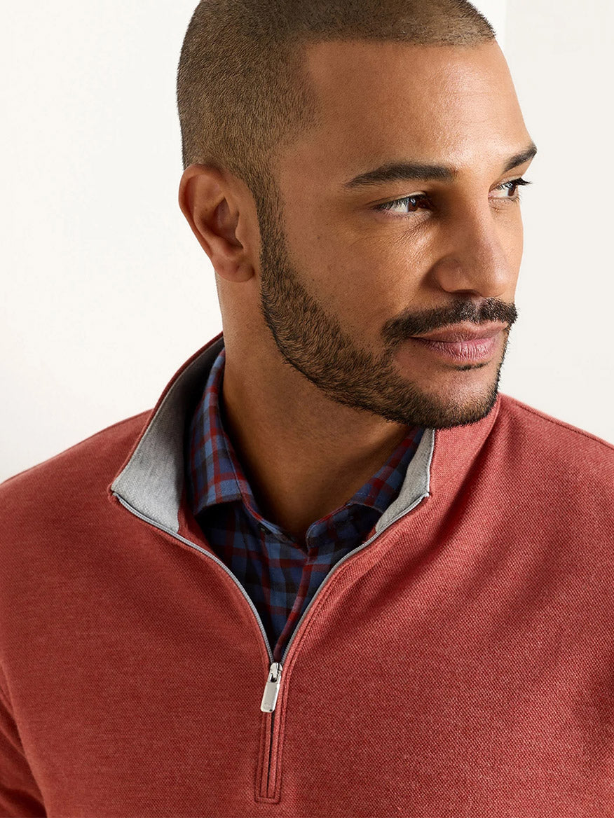 A man with closely-cropped hair and a beard wears the Peter Millar Crown Comfort Pullover in Burnt Orange, featuring four-way stretch, layered over a blue plaid shirt, as he looks to his right against a plain background.