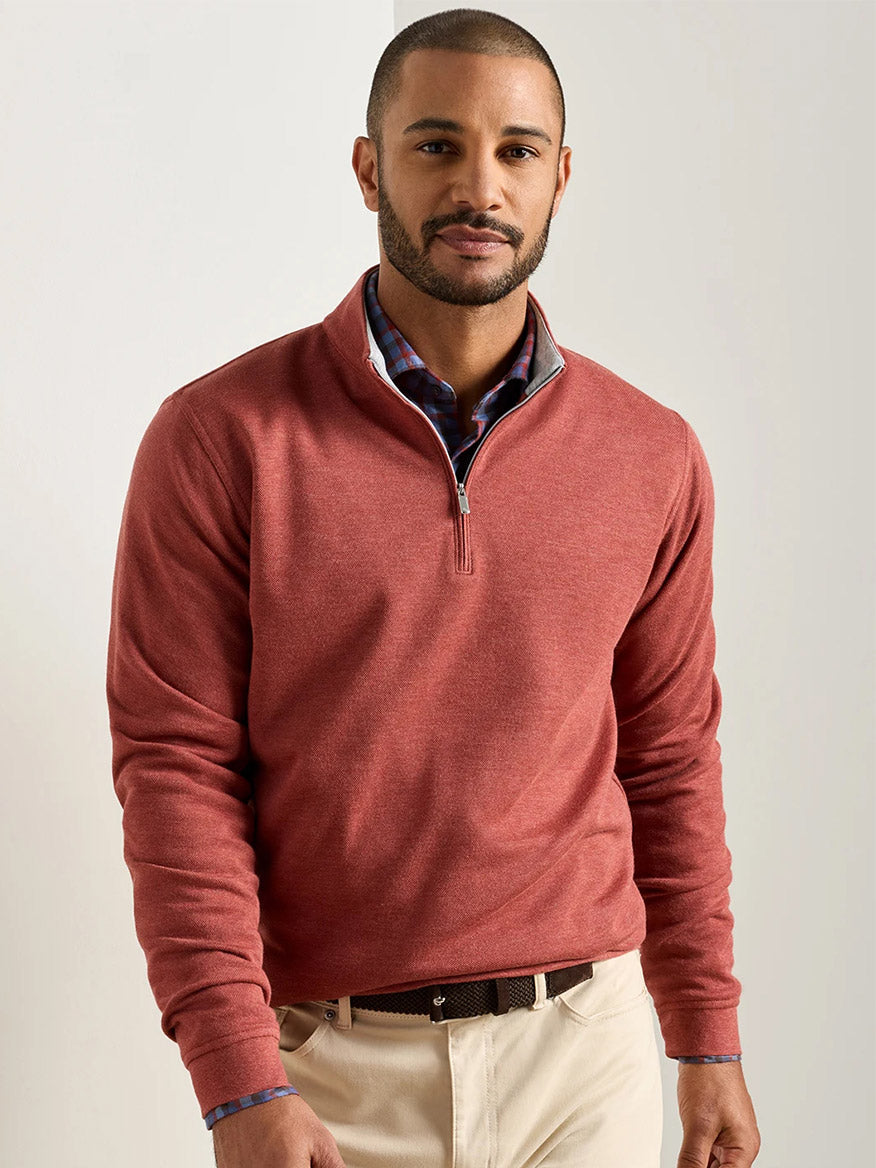 A man stands indoors wearing the Peter Millar Crown Comfort Pullover in Burnt Orange over a plaid shirt and beige pants, looking at the camera with a neutral expression.