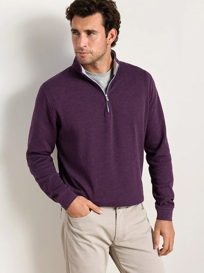 A man wearing the Peter Millar Crown Comfort Pullover in Grapeseed and light-colored pants stands against a plain light wall, looking to his left with one hand in his pocket. This four-way stretch quarter-zip delivers lightweight comfort and easy movement for any occasion.