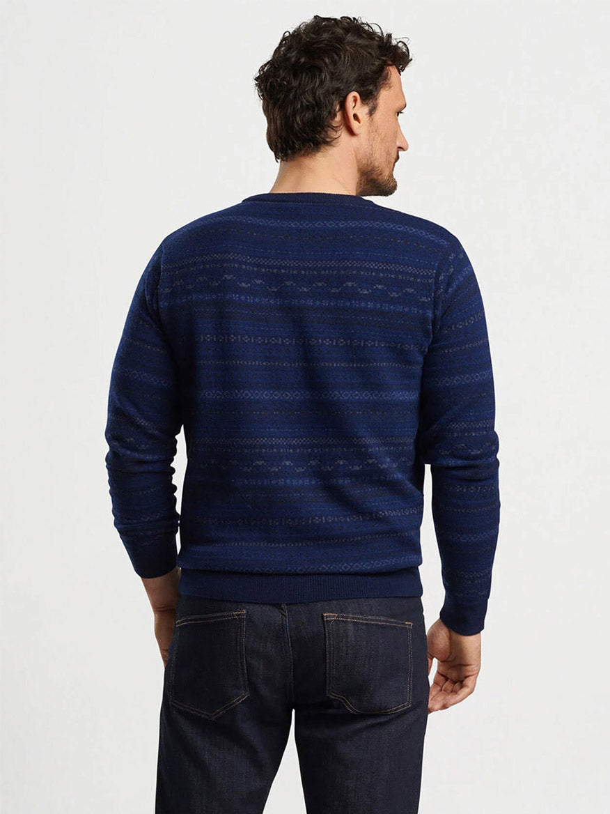 A man with curly hair stands with his back to the camera, wearing the Peter Millar Midnight Fairisle Crewneck Sweater in Navy, crafted from a cashmere blend, along with dark jeans.