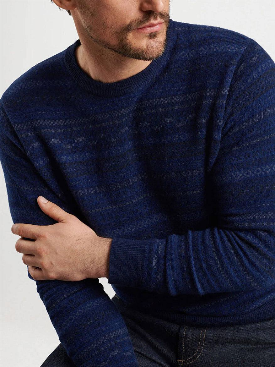 A person wearing a navy Peter Millar Midnight Fairisle Crewneck Sweater sits with their arm resting on their knee.