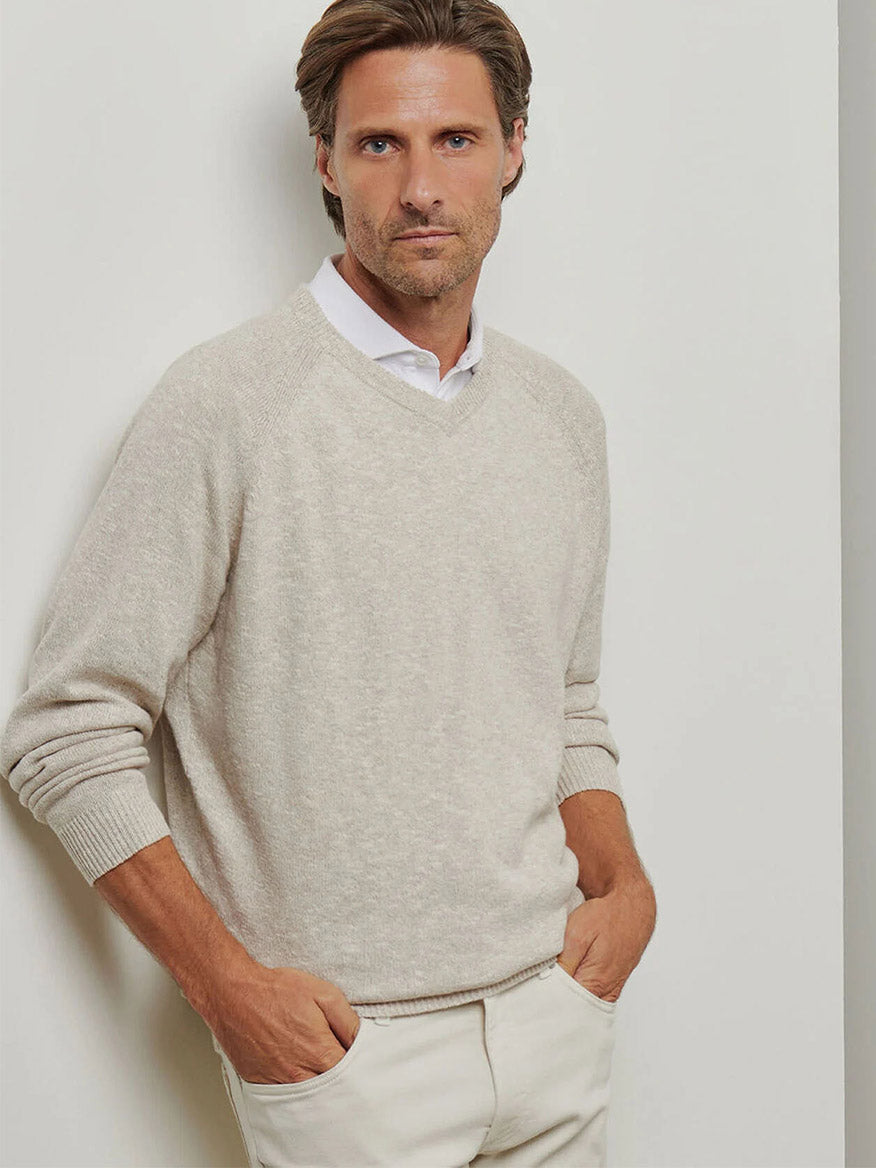 A man in a casual Peter Millar Montauk V-Neck Sweater in Palm Thatch, layered over a crisp white shirt, stands confidently with hands in pockets against a neutral backdrop.