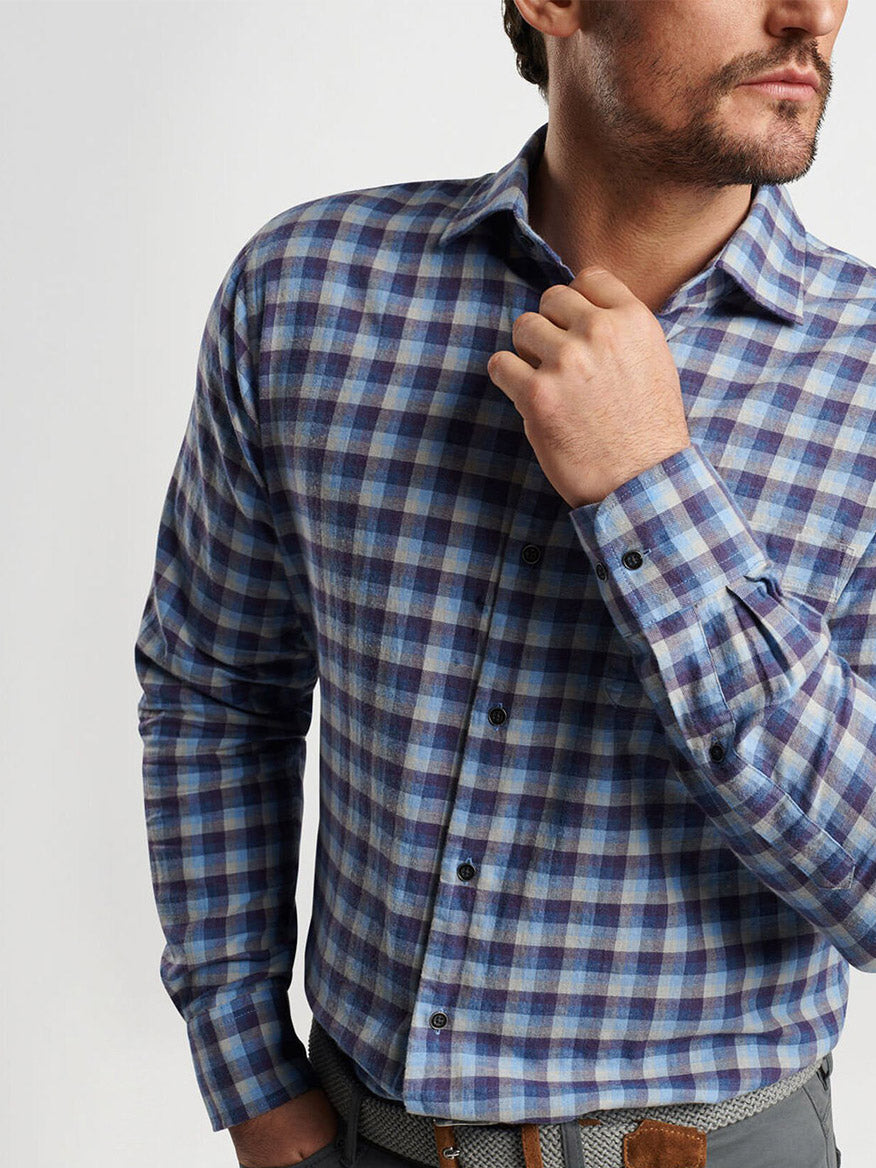 A person wearing the Peter Millar North Bay Cotton Sport Shirt in Ocean Blue, a breathable cotton twill shirt featuring a blue and gray plaid pattern, adjusts the collar to highlight its classic fit.