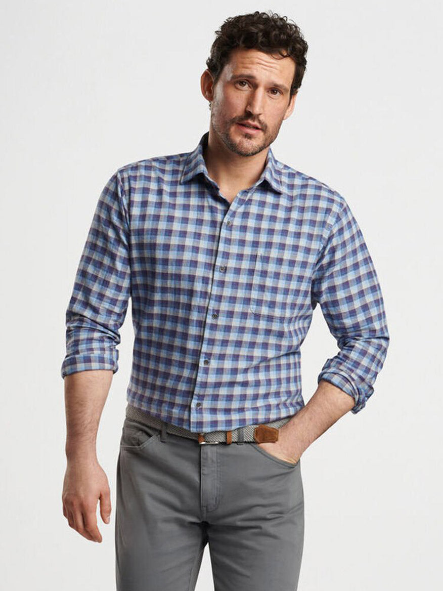 A man wearing the Peter Millar North Bay Cotton Sport Shirt in Ocean Blue, with its classic fit and checkered pattern, has the sleeves rolled up. Paired with gray pants and set against a plain background, he exudes a breathable sense of style.