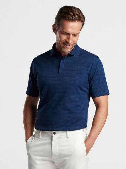 A man wearing a Peter Millar Pembroke Polo in Blue Pearl and white pants, looking downwards with a slight smile, against a light gray background.