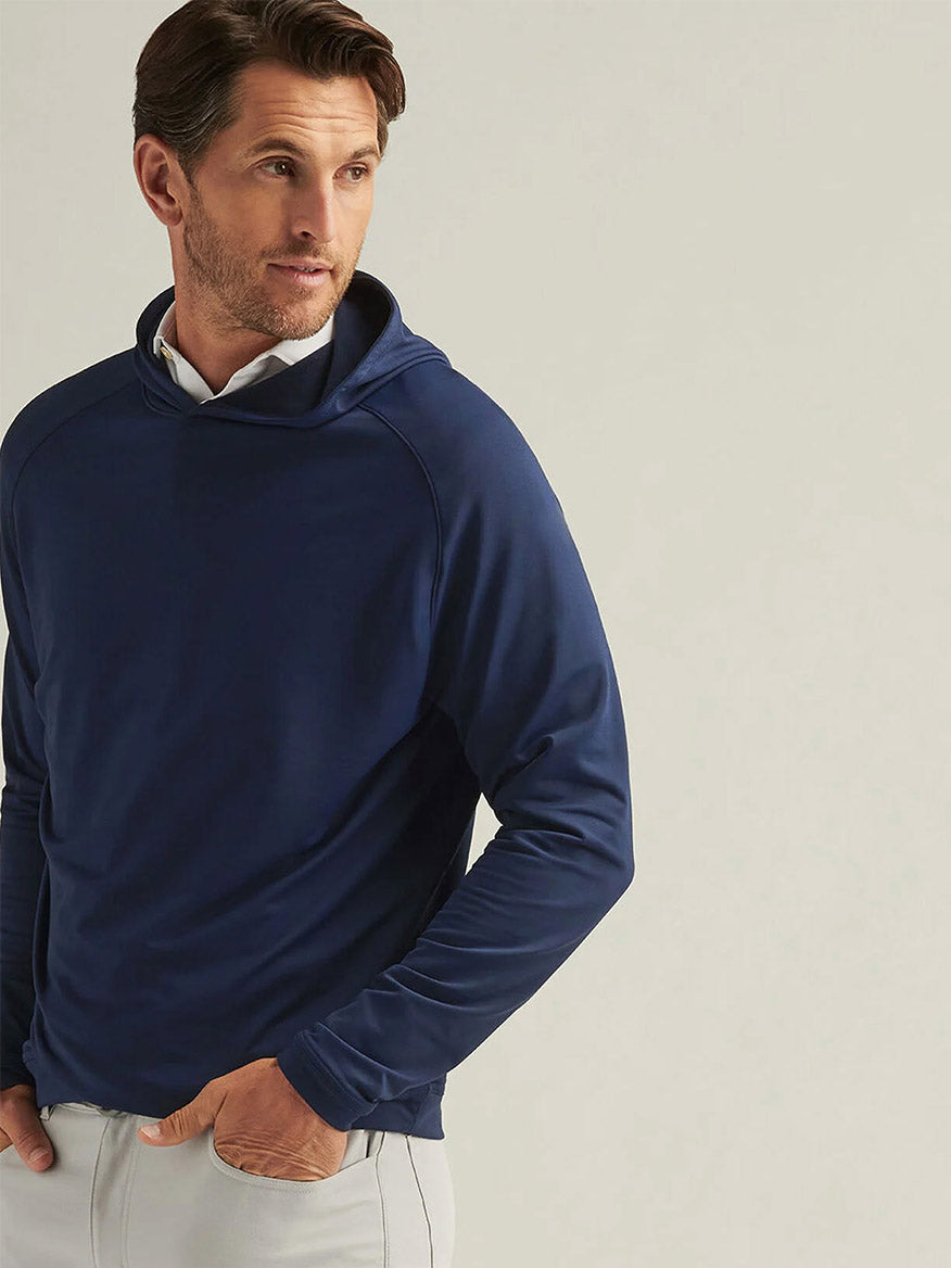 A man wearing the Peter Millar Pine Performance Hoodie in navy and light-colored pants stands with his hands in his pockets, looking to the side against a plain background.