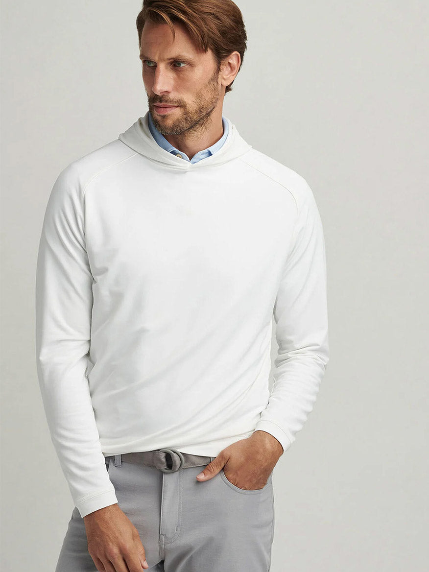 A man wearing the Peter Millar Pine Performance Hoodie in White and gray pants stands with one hand in his pocket against a plain background.