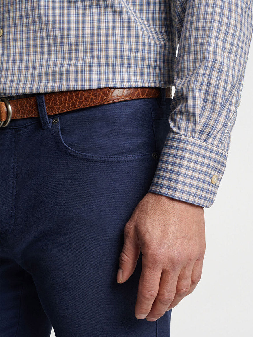 A person wearing a Peter Millar Prairie Performance Twill Sport Shirt in Galaxy tucked into dark blue pants with a brown leather belt. The person's left hand is resting by their side.