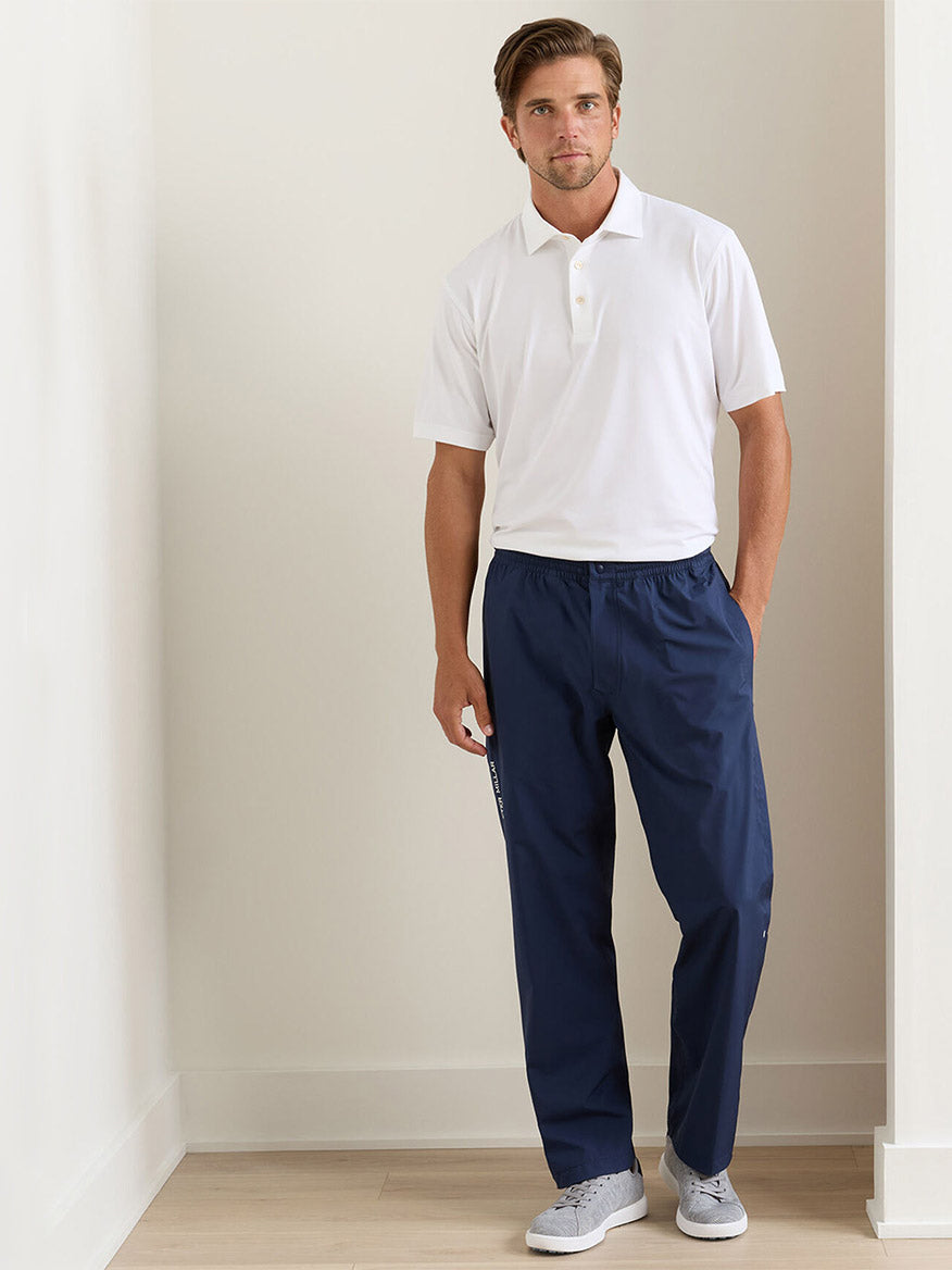 A man stands indoors against a plain white wall wearing a white polo shirt, gray sneakers, and Peter Millar Rain Walker Pant in Navy with one hand in his pocket.