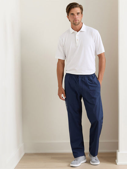 A man stands indoors against a plain white wall wearing a white polo shirt, gray sneakers, and Peter Millar Rain Walker Pant in Navy with one hand in his pocket.