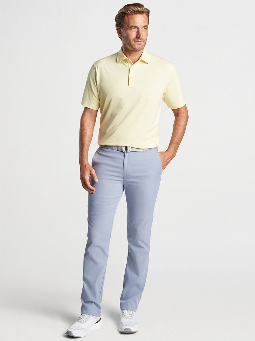 A man stands against a neutral background wearing a yellow polo shirt, Peter Millar Raleigh Performance Trouser in London, and white shoes.