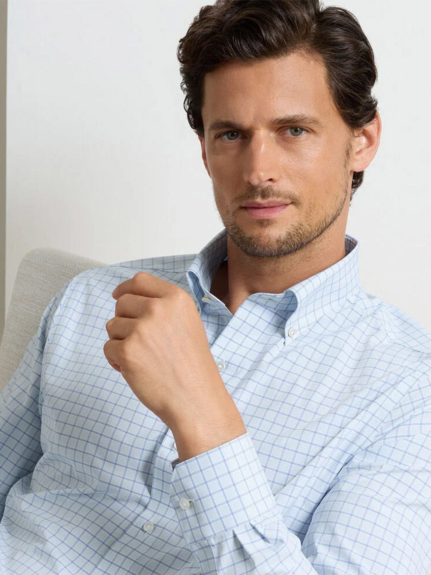 A man with short brown hair and a trimmed beard wears a Peter Millar Rampart Performance Poplin Sport Shirt in Blue Brisk, sitting and looking at the camera with a neutral expression.