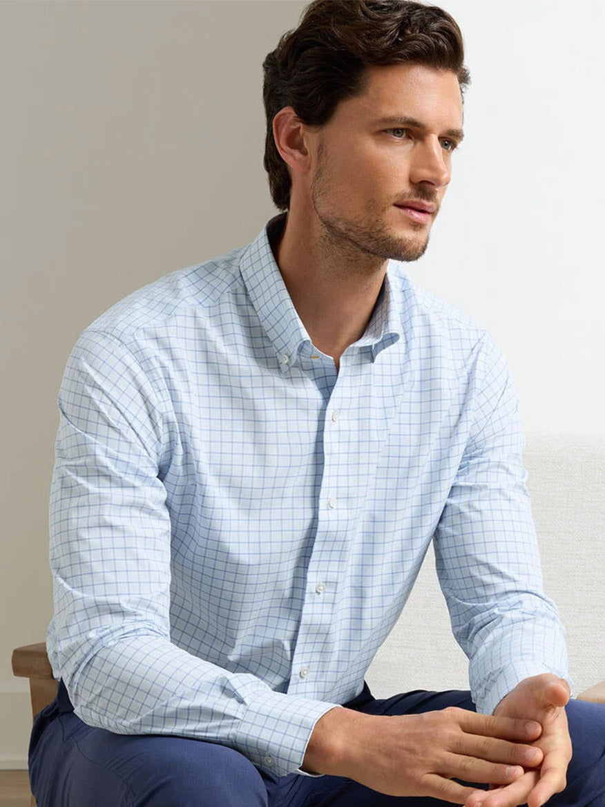 A man wears the Peter Millar Rampart Performance Poplin Sport Shirt in Blue Brisk and dark blue pants while seated on a light chair, looking to the side. The performance fabric ensures all-day comfort.