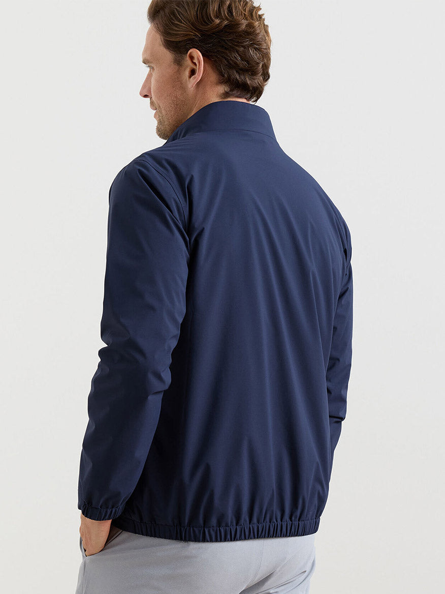 A man is seen from behind wearing the Peter Millar Ryder 3-in-1 Jacket in Navy, featuring a stand collar and elastic cuffs, set against a plain light background.