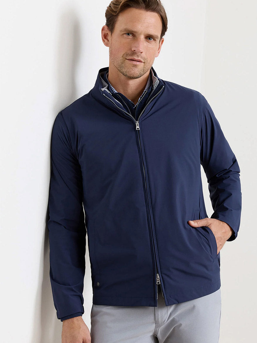 A man stands against a plain light-colored wall with one hand in his pocket, wearing the Peter Millar Ryder 3-in-1 Jacket in Navy over a collared shirt.