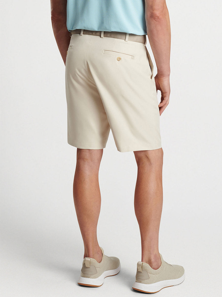 Man wearing beige Peter Millar Salem Performance Short in Stone and light-colored sneakers standing with hands in pockets.