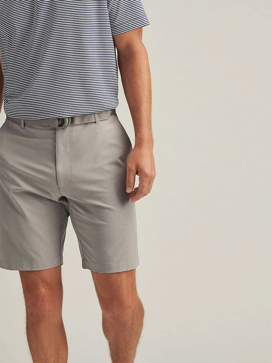 A person wearing light gray Peter Millar Shackleford Performance Hybrid Shorts in British Grey and a short-sleeve, striped polo, standing against a plain background.