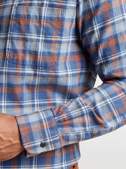 A close-up shows someone dressed in the Peter Millar Sherbrooke Cotton Sport Shirt in Ocean Blue, featuring a classic fit with blue, brown, and white plaid pattern. The focus is on the chest pocket and sleeve with buttoned cuff, underscoring the breathable cotton twill material.