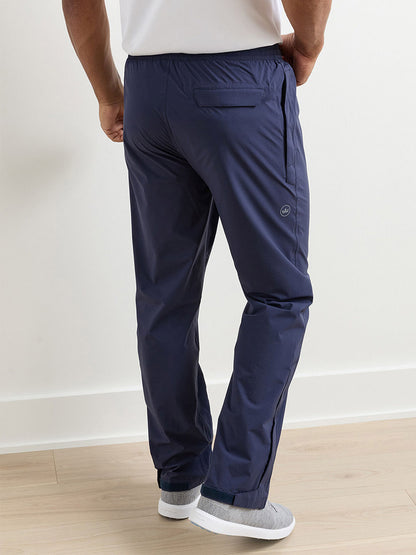 Someone stands with their back to the camera on a light wood floor, wearing navy Peter Millar Shield Pull-On Rain Pants and gray slip-on shoes.