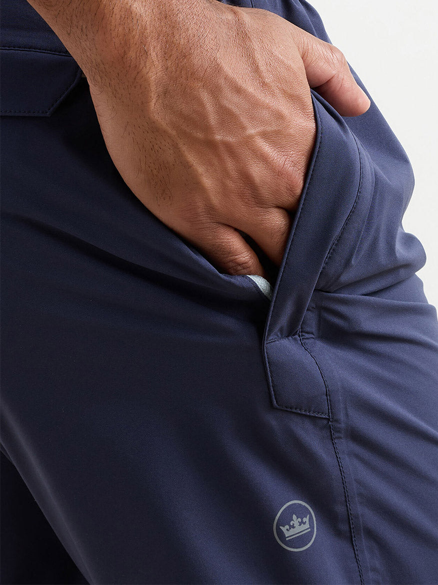 A close-up of a hand in the pocket of Peter Millar Shield Pull-On Rain Pant, featuring dark blue waterproof fabric, seam-sealed protection, and a small crown logo near the pocket seam.