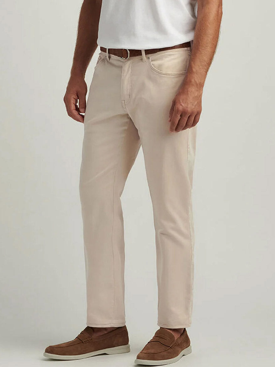 A man wears Peter Millar Signature Sateen Five-Pocket Pant in Sand, a white t-shirt, a brown belt, and brown loafers as he stands against a plain background.
