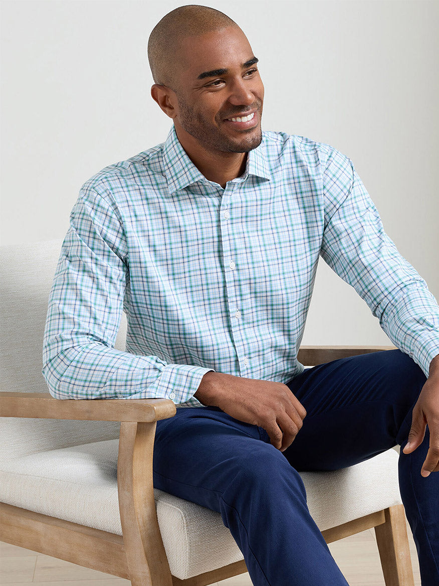 A man sits on a light chair, smiling and looking to the side, wearing dark blue pants and the Peter Millar Simon Crown Lite Cotton-Stretch Sport Shirt in Windsor Blue.