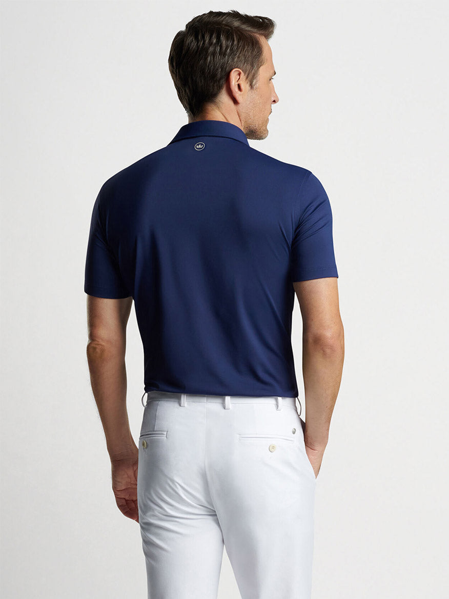 Man wearing a navy blue Peter Millar Solid Performance Jersey polo shirt and white pants viewed from the back.