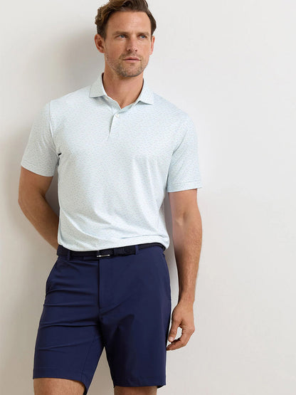 A man wearing a Peter Millar Soprano Waves Performance Jersey Polo in white and dark blue shorts stands against a plain light background.