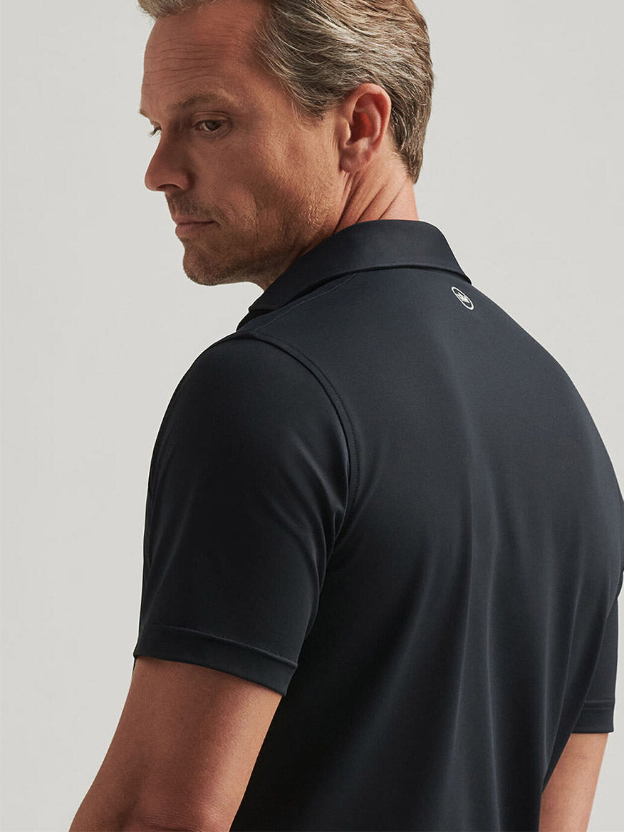 A man wearing a Peter Millar Soul Performance Mesh Polo in Black, featuring mother-of-pearl buttons, glances over his shoulder against a plain background.