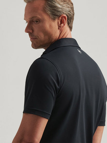 A man wearing a Peter Millar Soul Performance Mesh Polo in Black, featuring mother-of-pearl buttons, glances over his shoulder against a plain background.