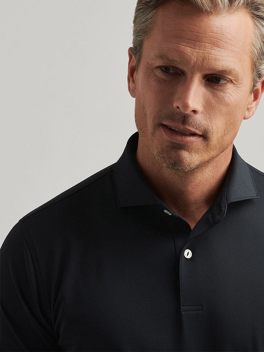 A man wears the Peter Millar Soul Performance Mesh Polo in Black, featuring mother-of-pearl buttons, as he looks slightly downward against a neutral background.