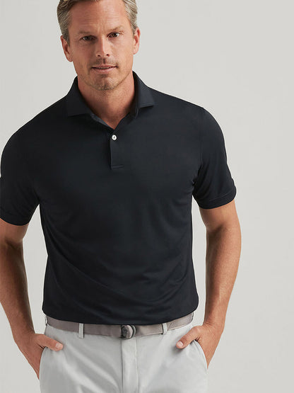 A man in a Peter Millar Soul Performance Mesh Polo in black, highlighted by mother-of-pearl buttons, paired with light gray pants, stands against a plain background with a neutral expression.