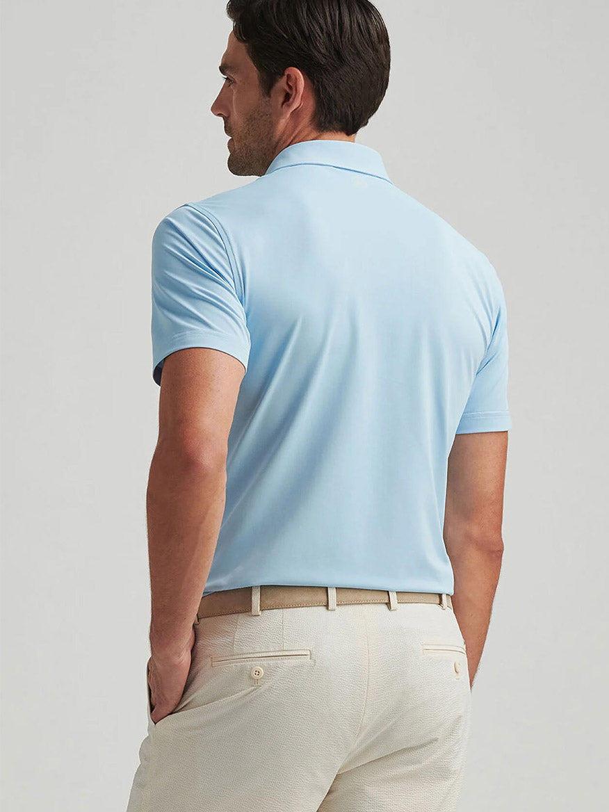 A man faces away, wearing the Peter Millar Soul Performance Mesh Polo in Blue Frost and beige pants with a belt against a plain background.
