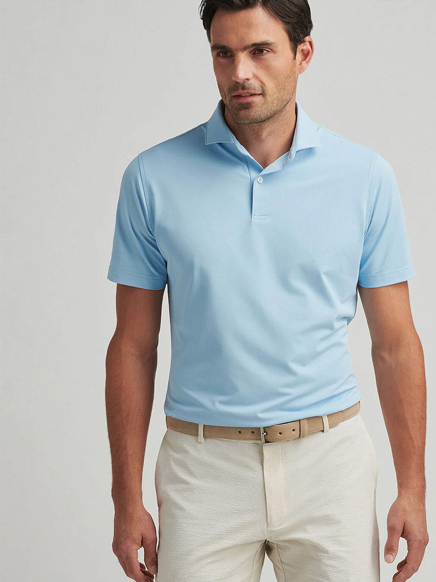 Man wearing a Peter Millar Soul Performance Mesh Polo in Blue Frost with four-way stretch, beige belt, and light-colored pants, standing against a plain light background.