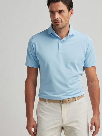 Man wearing a Peter Millar Soul Performance Mesh Polo in Blue Frost with four-way stretch, beige belt, and light-colored pants, standing against a plain light background.