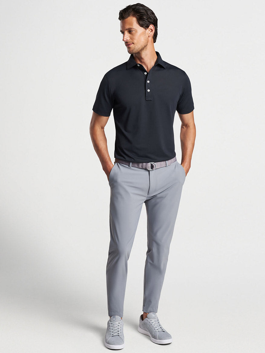 Man posing in a Peter Millar Soul Performance Mesh Polo in Black and grey trousers with a belt, paired with light grey sneakers.