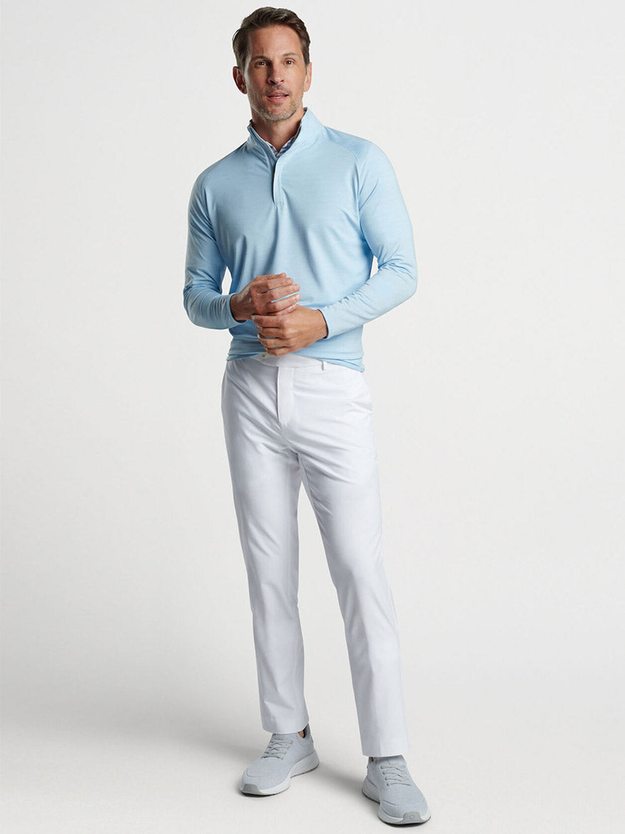 Man wearing a Peter Millar Stealth Performance Quarter-Zip in Blue Frost with a zip collar and white trousers, posing with hands clasped in front of him.