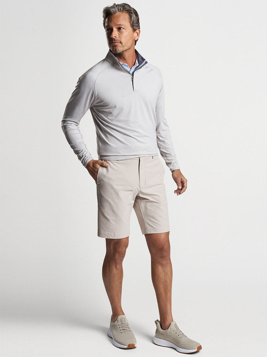 A man posing in a smart-casual outfit with a Peter Millar Stealth Performance Quarter-Zip in British Grey made from wicking fabric, beige shorts, and beige sneakers.