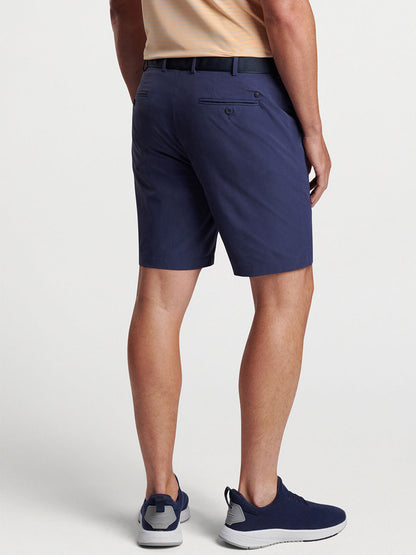 A person standing side-on wearing Peter Millar Surge Performance Short in Navy, a belt, and sporty blue sneakers.